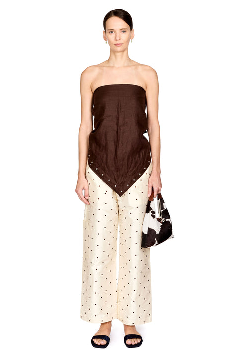 A model stands against a white studio background wearing a strapless dark brown top with a draped, asymmetrical hem and matching dark polka-dot wide-leg pants. She is holding a black and white patterned handbag and wearing black slide sandals.