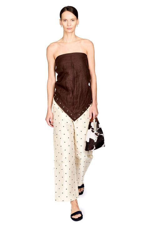 A model wearing a dark brown strapless top with a draped, asymmetrical hem and cream wide-leg pants with black polka dots, holding a matching brown and black patterned handbag against a plain white background.