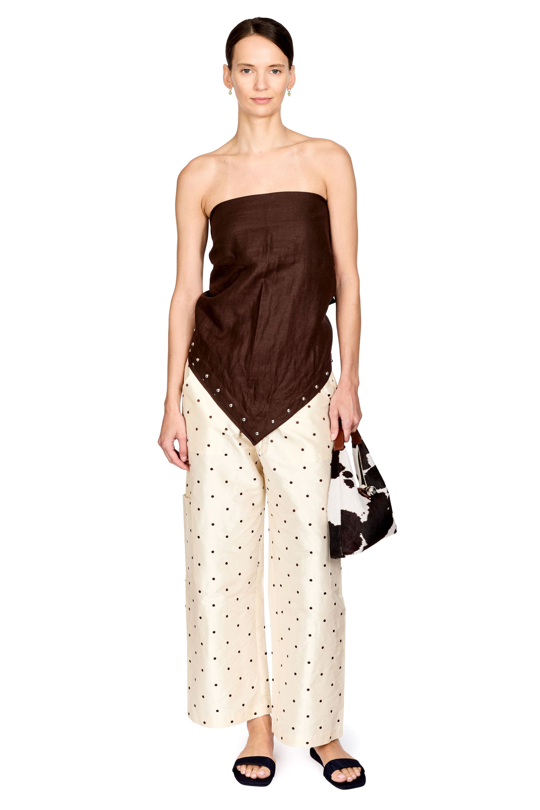 A model stands against a white background wearing a dark brown strapless top with a draped triangular front panel, paired with cream wide-leg pants featuring small black polka dots, black slide sandals, and holding a black-and-white patterned handbag.