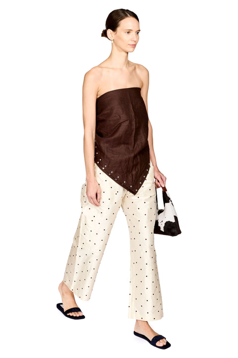 A model wearing a dark brown strapless top with silver studs and cream wide-leg pants with black polka dots, holding a black and white handbag against a plain white background.