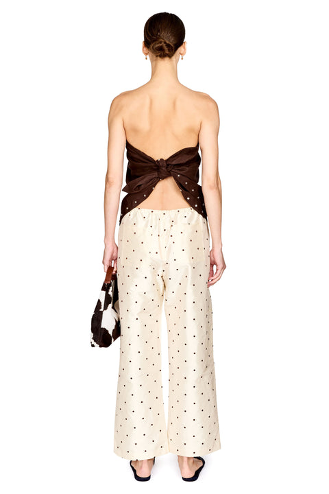A model shown from the back wearing a strapless dark brown top with a large bow detail and cream wide-leg pants with small black polka dots, holding a small black and white handbag against a plain white background.