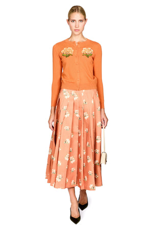 A model stands against a white background wearing a two-piece outfit consisting of a burnt orange long-sleeve button-up top with floral embroidery on the chest and a matching peach-orange pleated midi skirt with a white and green floral print. She is holding a small beige handbag and wearing black pointed-toe heels.