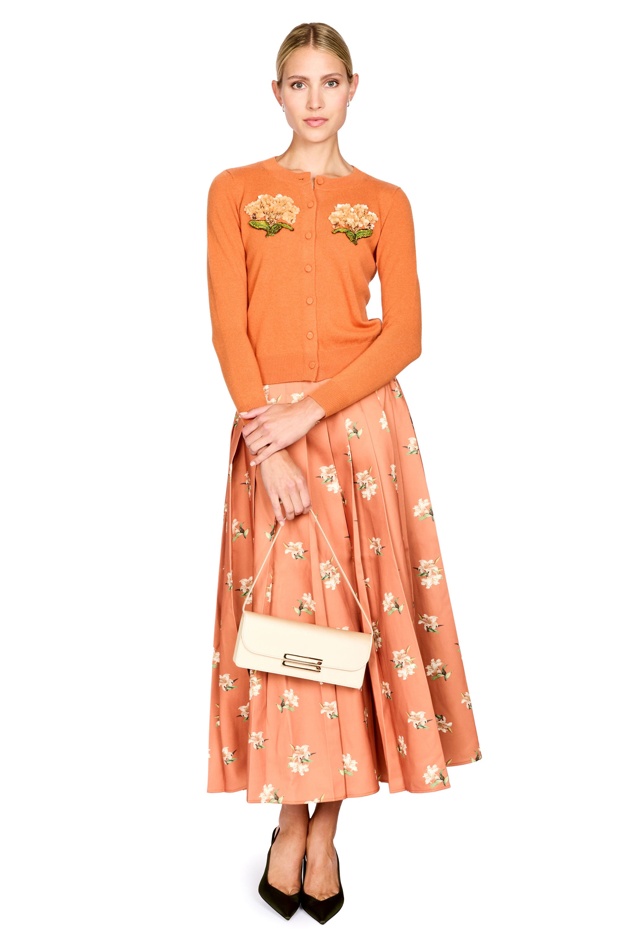 A woman modeling a two-piece outfit consisting of a short-sleeve orange knit cardigan with floral embroidery on the chest and a matching orange floral midi skirt. She is holding a cream-colored handbag and wearing black pointed-toe heels, set against a plain white background.