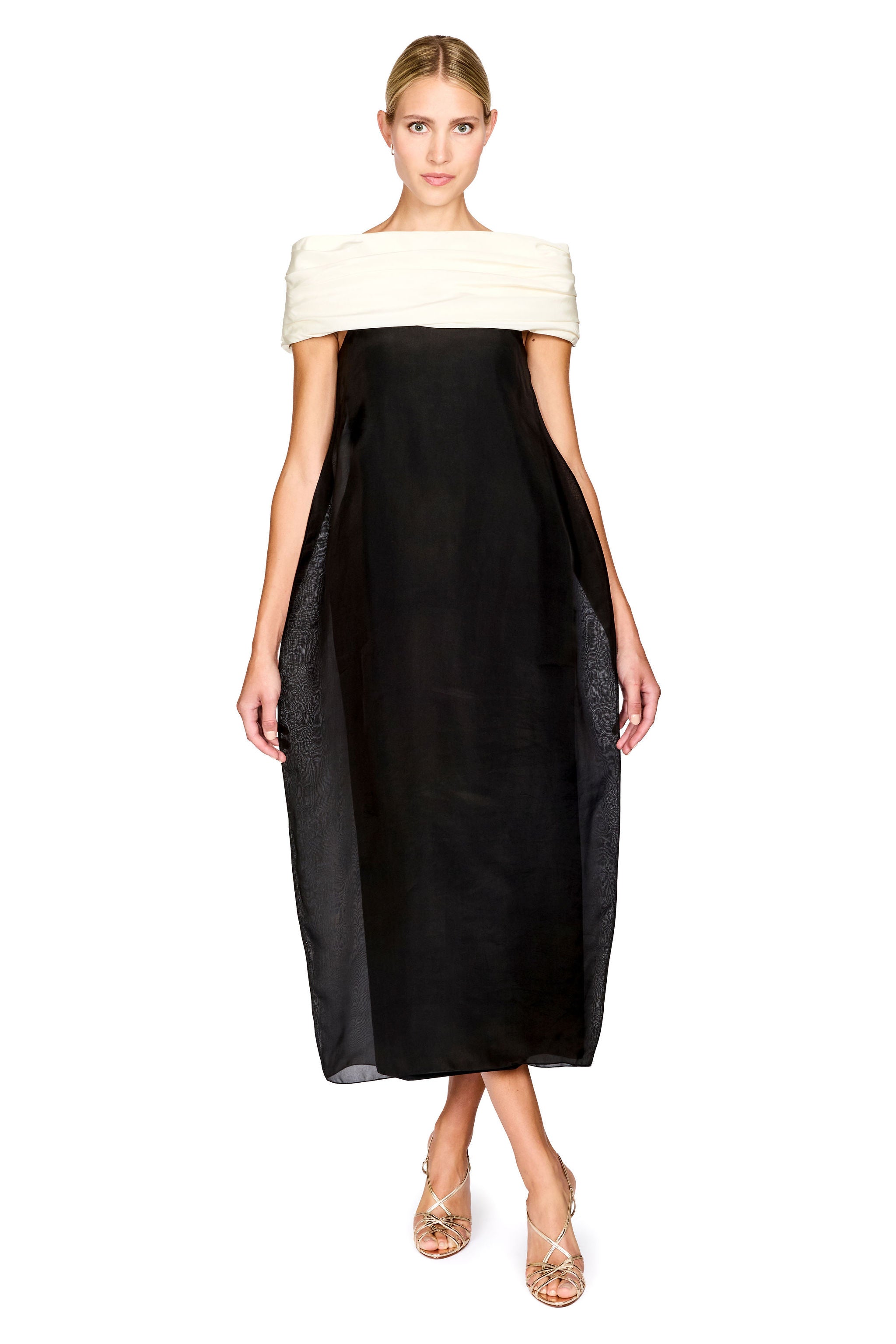 A model wearing a black midi dress with a wide off-the-shoulder cream strap and sheer black side panels, paired with clear high-heeled sandals, against a plain white background.