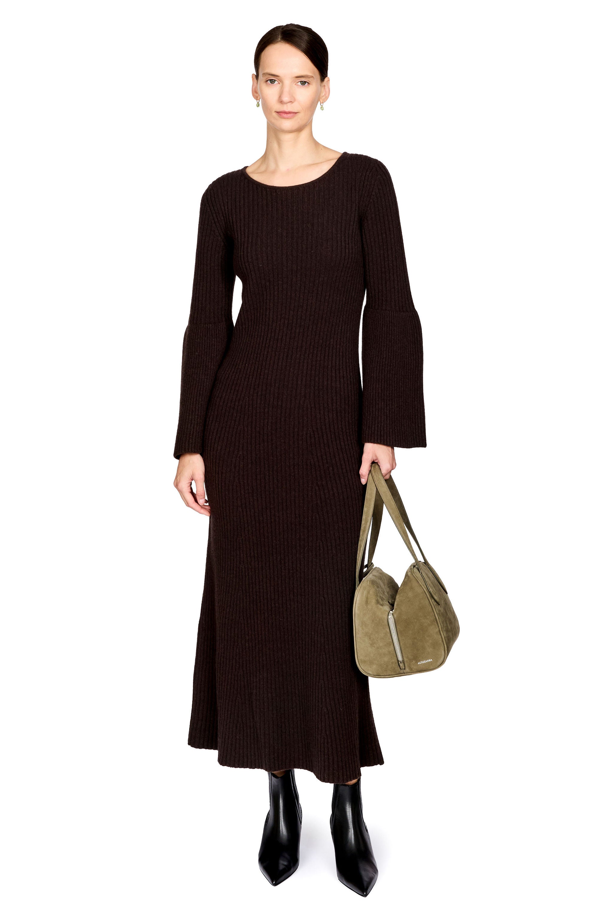 A model stands against a white background wearing a dark brown ribbed knit long-sleeve maxi dress, black pointed boots, and holding a taupe suede shoulder bag.