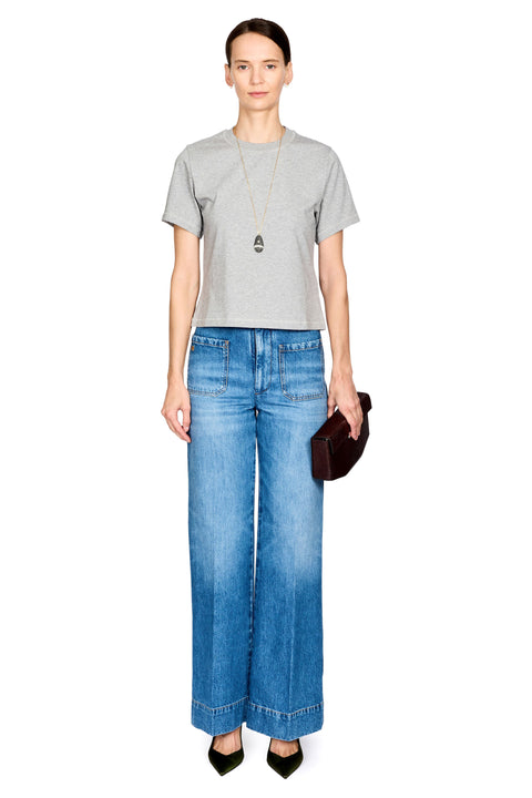 A model stands against a white background wearing a light gray short-sleeve t-shirt, wide-leg blue denim jeans, a long silver necklace, and black pointed shoes, while holding a dark brown clutch.