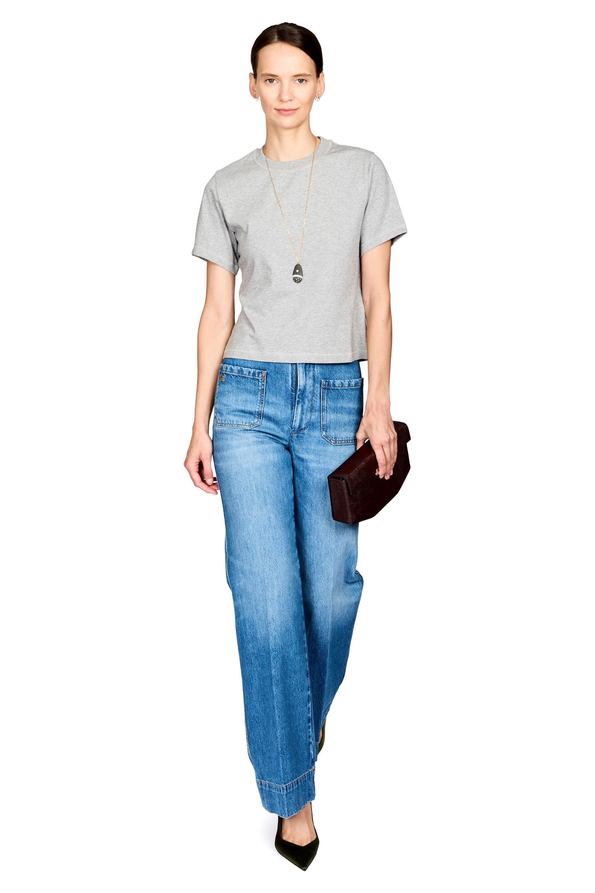 A woman modeling a light gray short-sleeve t-shirt, blue wide-leg jeans, a long silver pendant necklace, and black pointed-toe heels, while holding a dark brown leather clutch against a plain white background.