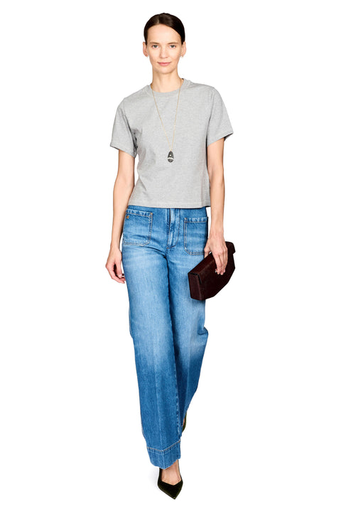 A woman modeling a light gray short-sleeve t-shirt, blue wide-leg jeans, a long silver pendant necklace, and black pointed-toe heels, while holding a dark brown leather clutch against a plain white background.
