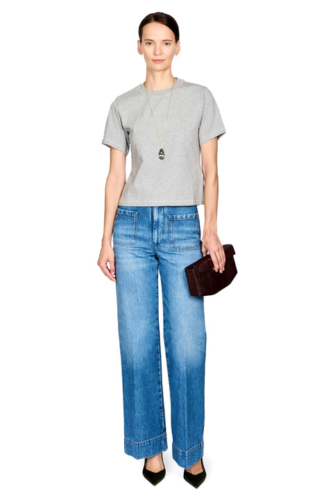 A model stands against a white background wearing a light gray short-sleeve t-shirt, blue wide-leg jeans, a long silver necklace, and holding a dark brown clutch.