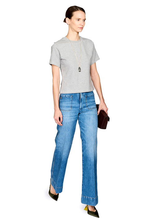 A model wearing a light gray short-sleeve t-shirt, blue wide-leg jeans, and dark green pointed-toe flats, holding a dark brown clutch against a plain white background.