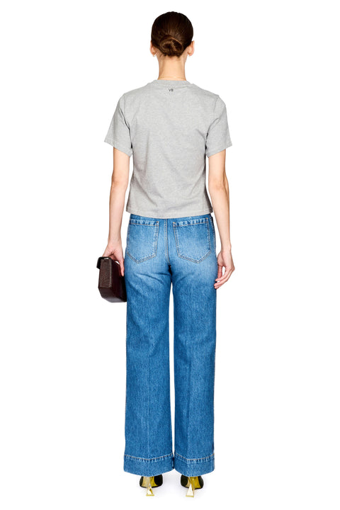 A model shown from the back wearing a light gray short-sleeve t-shirt, wide-leg blue denim jeans, and gold high heels, holding a small dark brown clutch against a plain white background.