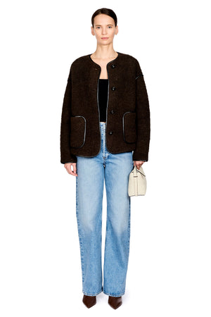 A model stands against a white studio background wearing a dark brown fuzzy cropped jacket over a black top, light blue straight-leg jeans, and brown pointed shoes, while holding a small beige handbag.