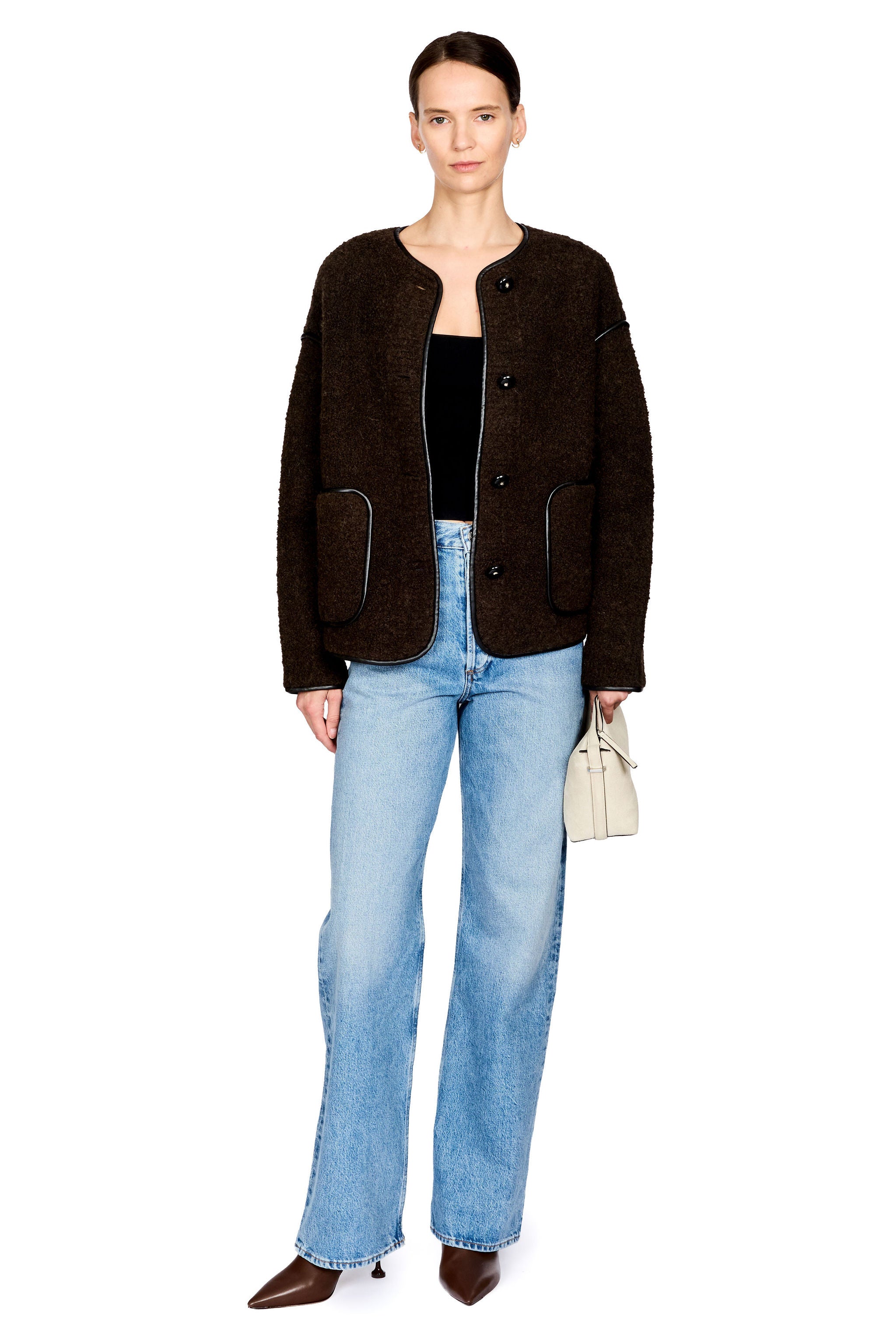 A model stands against a white studio background wearing a dark brown textured cropped jacket over a black top, light-wash blue jeans, brown pointed-toe heels, and holding a small beige handbag.