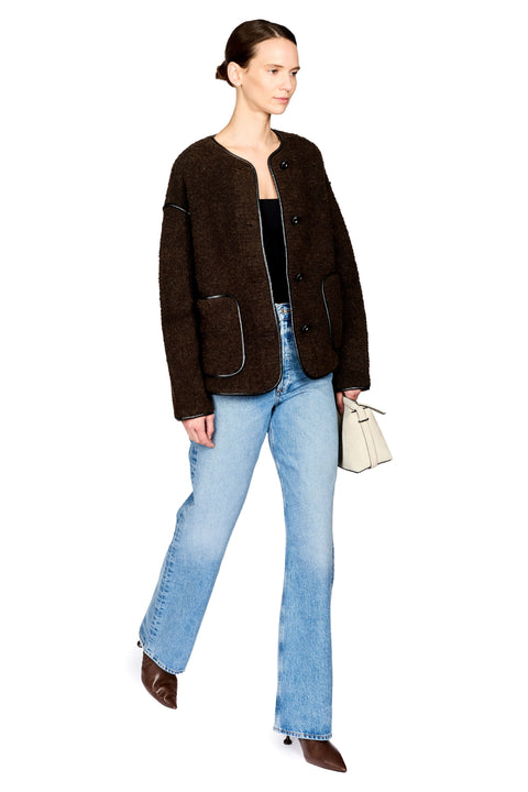 A model wearing a dark brown textured bouclé jacket with contrast piping, light-wash blue jeans, and brown pointed-toe boots, holding a small cream handbag against a plain white background.