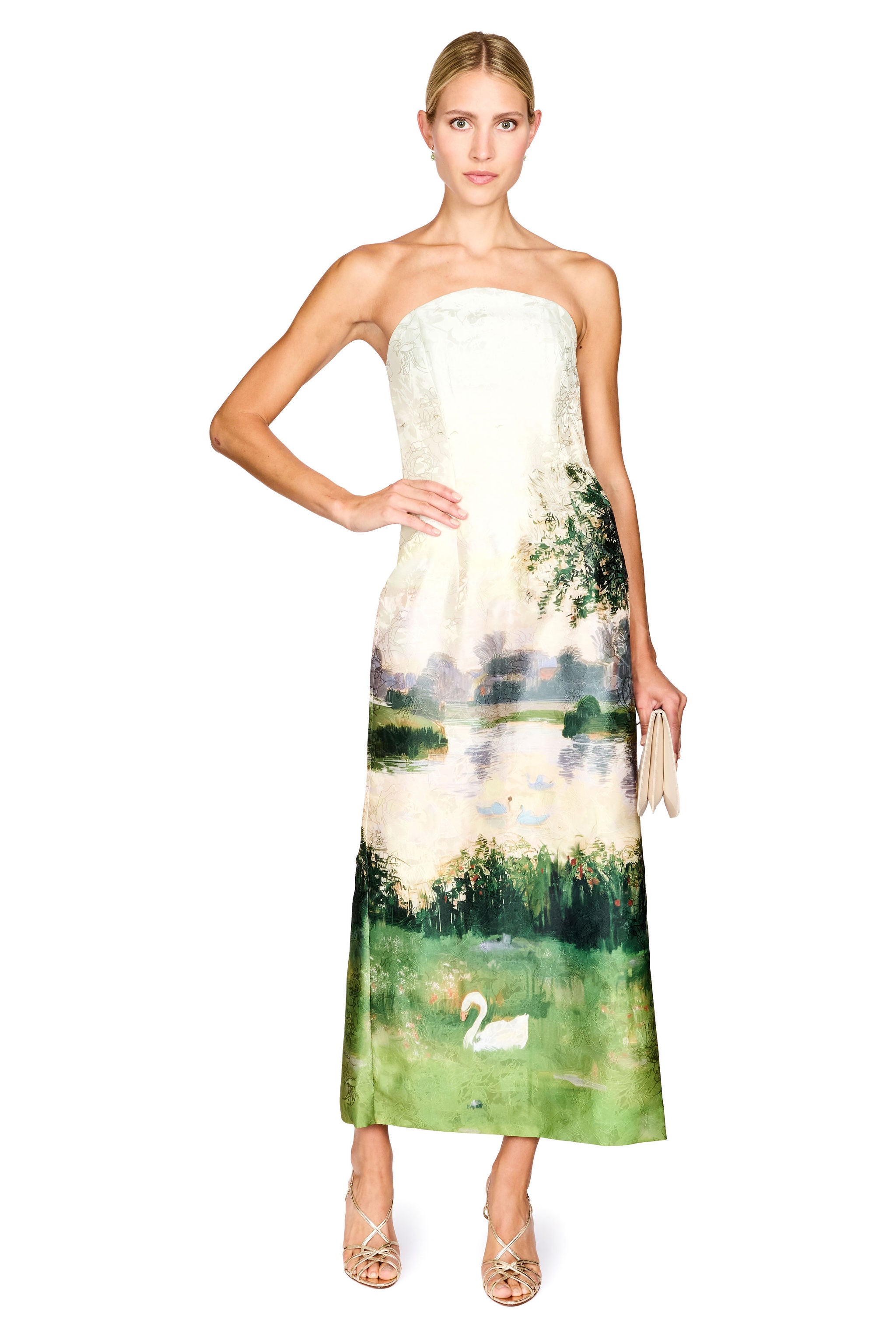A woman modeling a strapless maxi dress with a white lace bodice and a green landscape print featuring a lake, trees, and a swan, set against a plain white background.