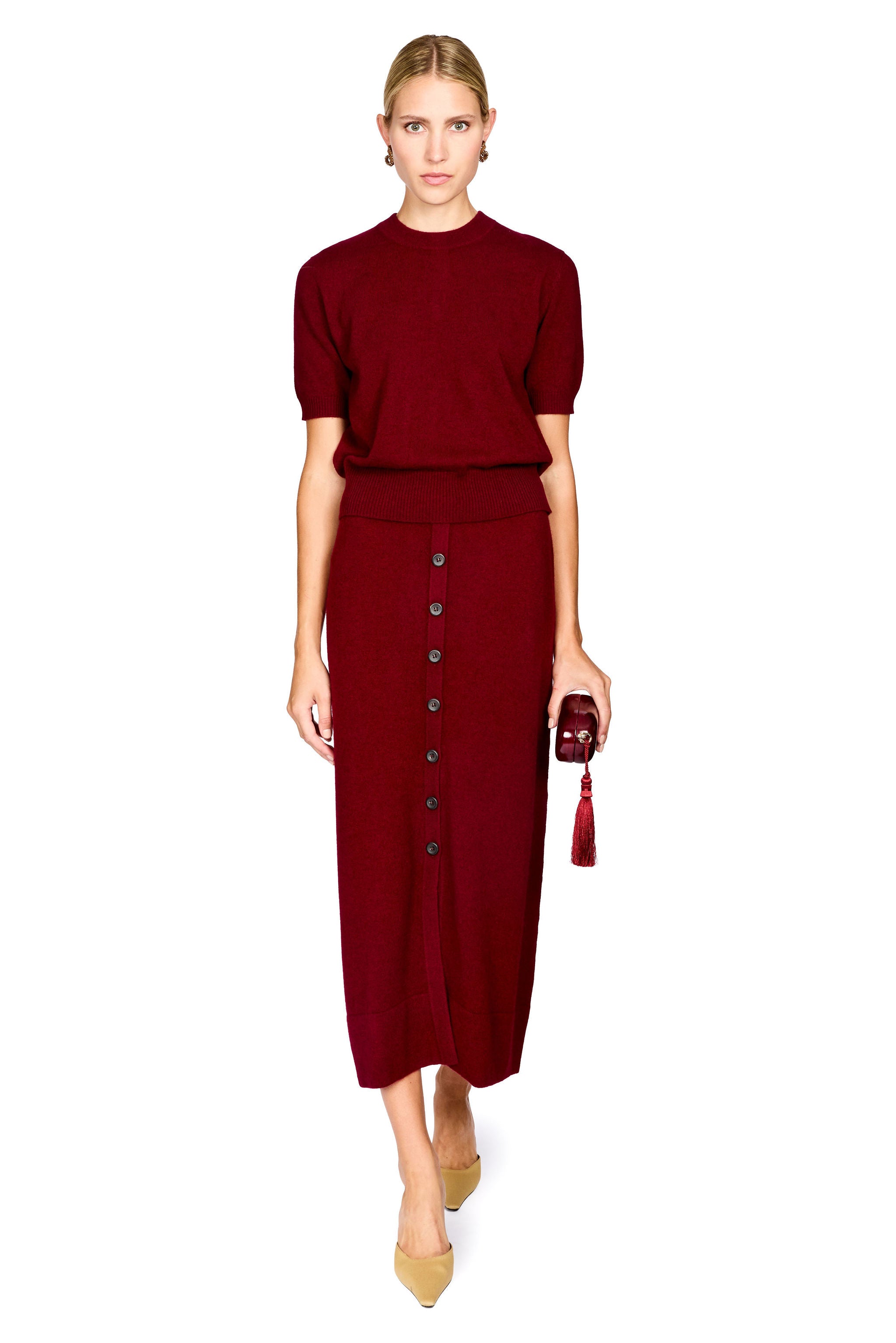 A woman modeling a matching burgundy knit two-piece set consisting of a short-sleeve mock-neck top and a high-waisted midi skirt with a button-front detail, paired with tan pointed-toe heels and a small matching clutch, on a solid white background.