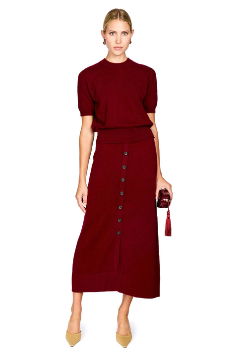 A woman modeling a matching burgundy knit short-sleeve top and midi skirt set with a button-front detail on the skirt, paired with beige pointed-toe heels and holding a small dark tassel bag, against a plain white background.