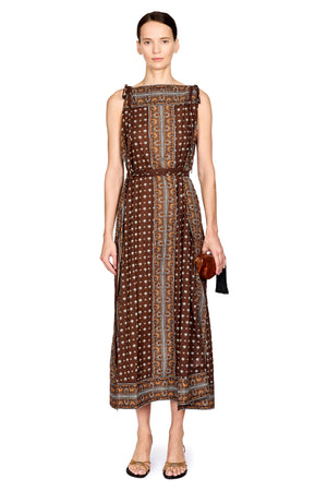 A woman modeling a sleeveless midi dress with a brown base, white polka dots, and vertical geometric patterns in blue, beige, and brown. She is standing against a solid white background.