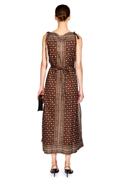 A woman viewed from the back wearing a sleeveless brown midi dress with a white and gold geometric pattern, tied at the shoulders, and paired with black heels against a white background.