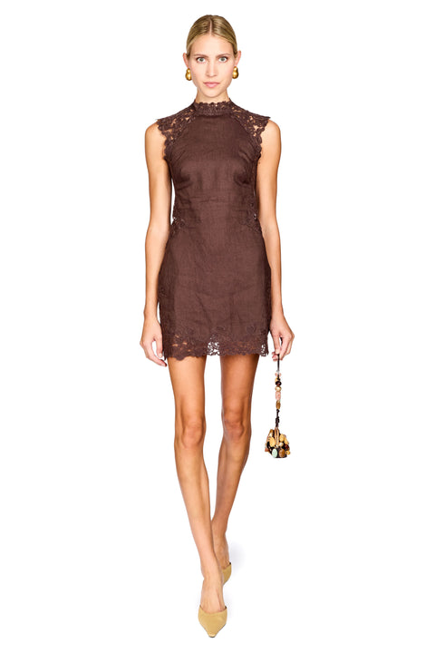 A model wearing a sleeveless, high-neck brown lace mini dress, holding a small patterned handbag, set against a plain white background.