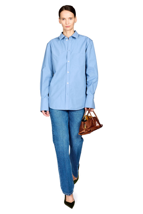 A woman modeling a light blue long-sleeve button-up shirt with blue jeans and black pointed shoes, holding a brown leather handbag against a plain white background.