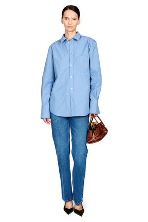 A woman modeling a light blue long-sleeve button-up shirt and blue jeans, holding a brown leather handbag against a plain white background.