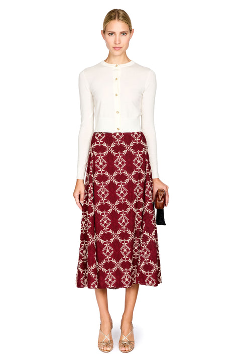 A woman modeling a white long-sleeve knit top and a burgundy midi skirt with a cream geometric trellis pattern, holding a small black clutch against a plain white background.