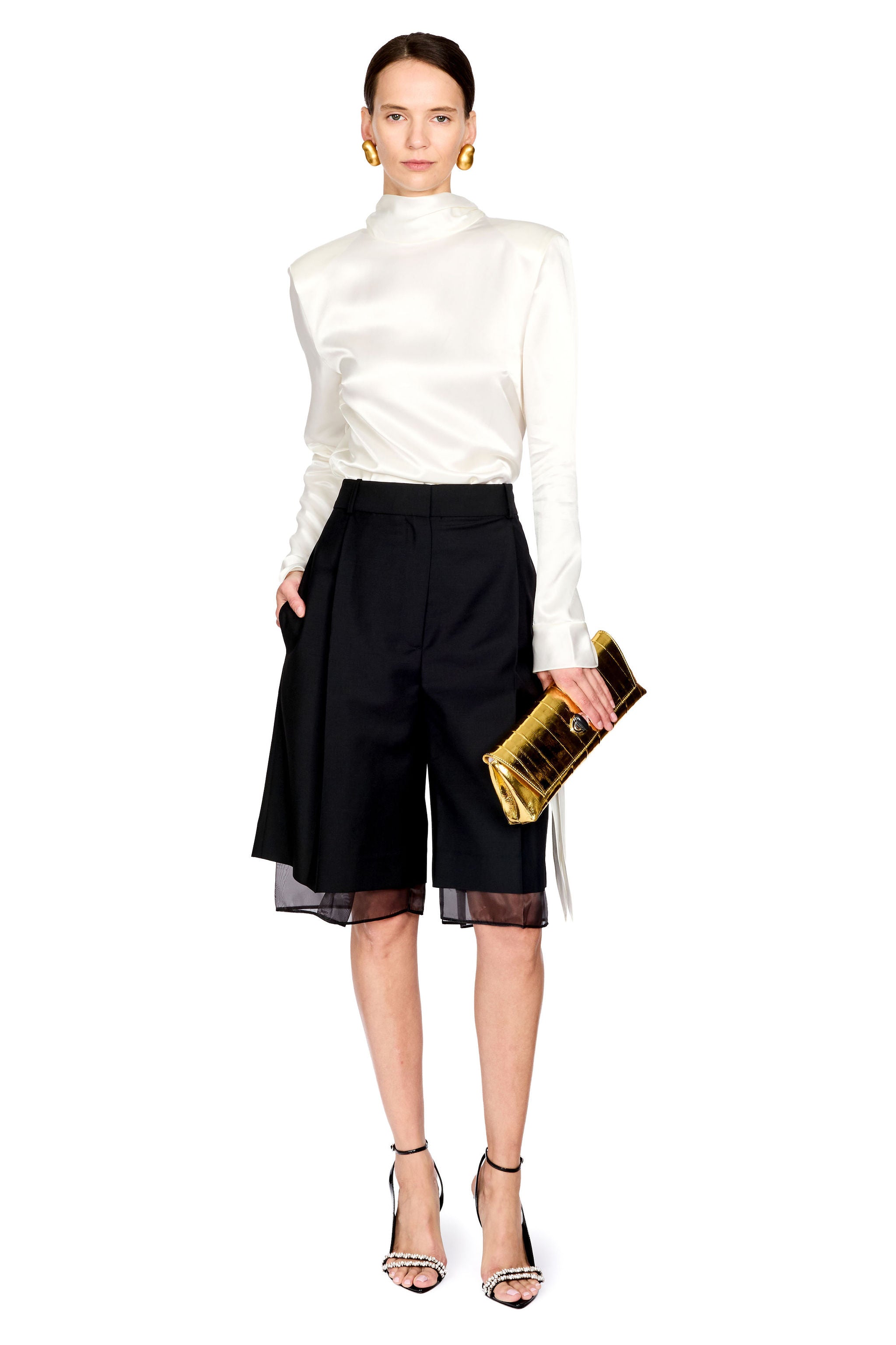 A model stands against a white studio background wearing a white satin mock-neck long-sleeve top, black pleated shorts with sheer black mesh hem, holding a gold clutch, and wearing gold hoop earrings and black high-heeled sandals.