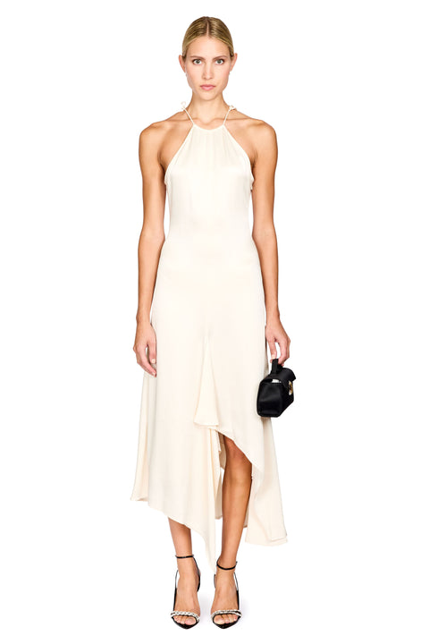 A woman modeling a sleeveless halter-neck midi dress in warm off-white/cream with an asymmetrical draped hem. She is standing against a plain white studio background, holding a small black handbag and wearing black strappy heels.