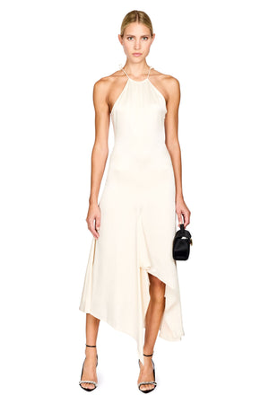 A woman modeling a cream-colored halter-neck midi dress with an asymmetrical draped hem, paired with black high-heeled sandals and a small black handbag, set against a plain white background.