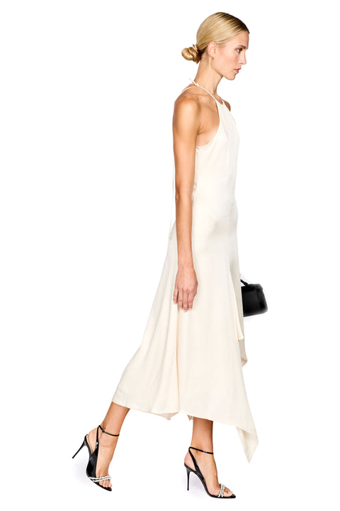 A woman modeling a sleeveless, high-neck, midi-length cream dress with an asymmetrical hem, paired with black strappy heels and a small black handbag, shown against a plain white background.