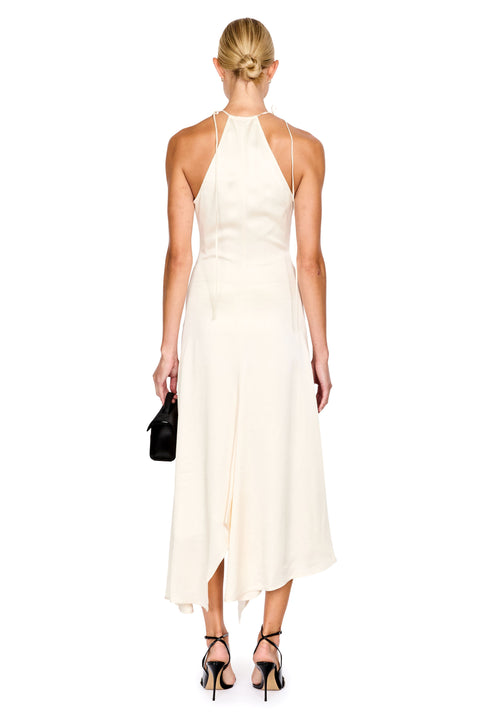 A woman shown from the back wearing a sleeveless cream halter midi dress with thin straps, a wrap-style skirt, and an asymmetrical hem. She is standing against a plain white background and holding a small black handbag.