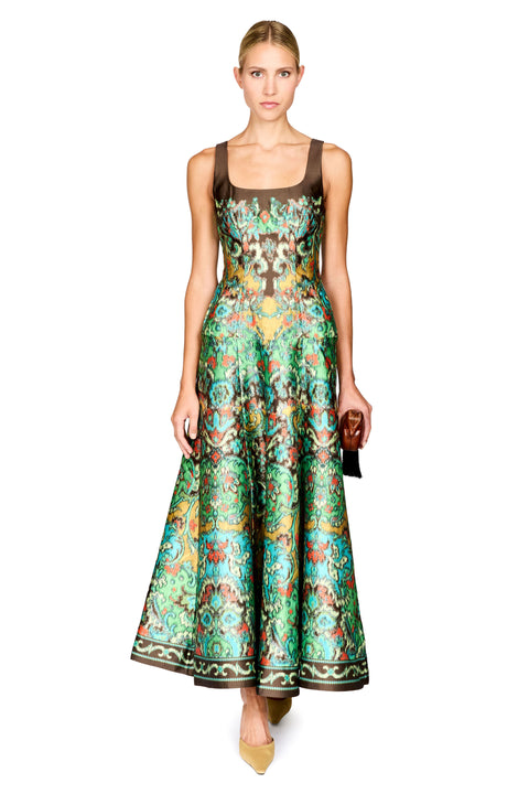 A model wearing a sleeveless, floor-length A-line gown with a brown bodice and a vibrant green, blue, and gold patterned skirt, holding a small brown handbag against a solid white background.