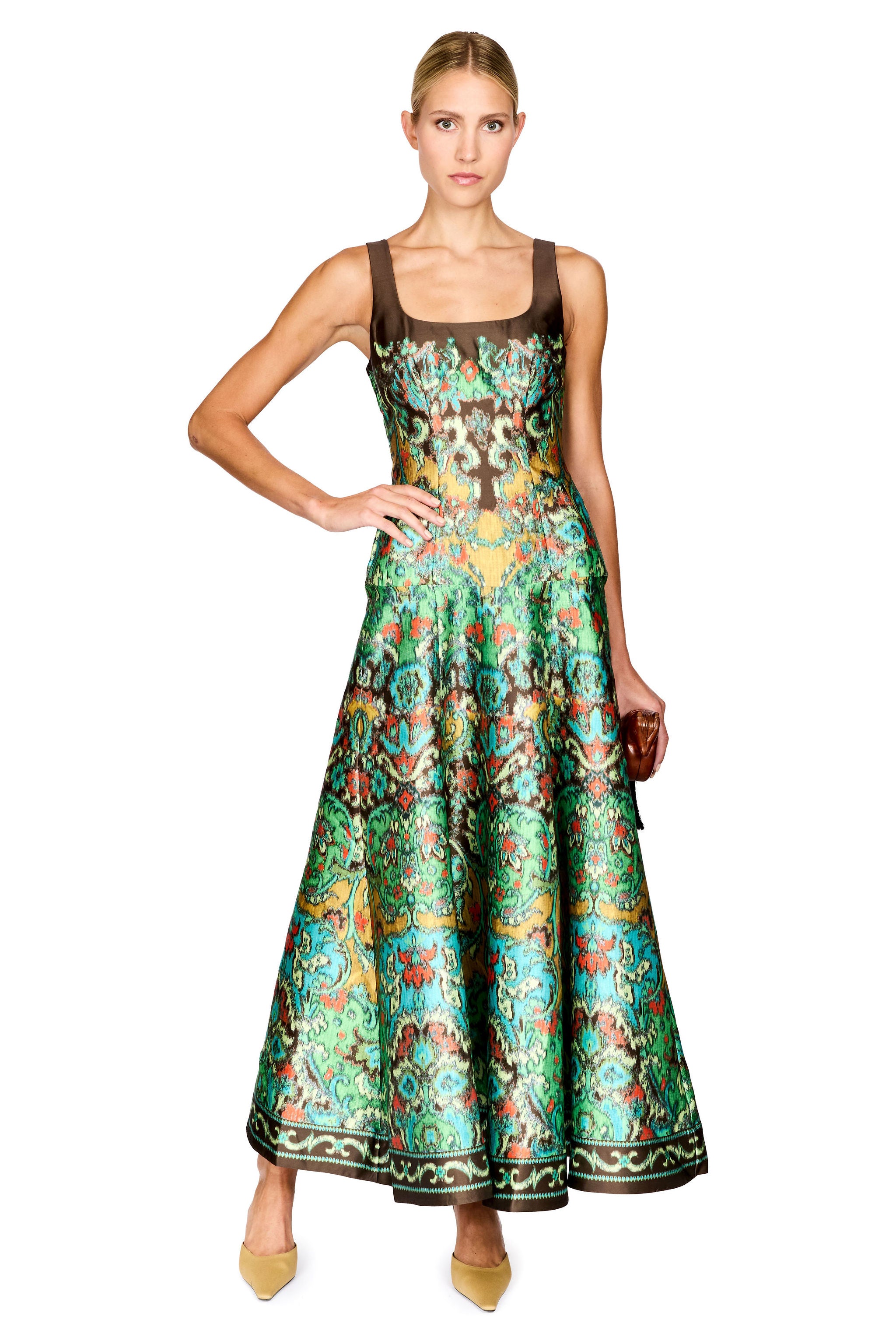 A woman modeling a sleeveless, floor-length A-line dress with a brown bodice and a vibrant green, blue, and gold patterned skirt, set against a plain white background.
