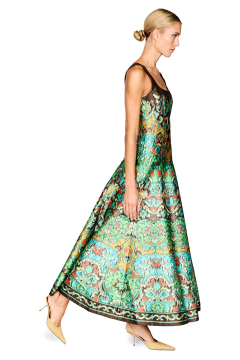 A woman modeling a sleeveless, floor-length maxi dress with a vibrant green, teal, and gold ornate pattern, paired with beige pointed-toe heels, set against a plain white background.