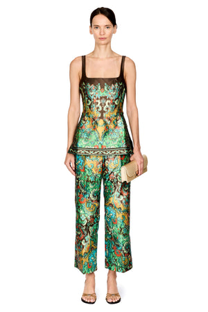 A model stands against a white background wearing a matching two-piece set consisting of a sleeveless top and wide-leg pants in a vibrant green, gold, and red patterned silk-like fabric. The set features a sheer brown mesh underlayer at the neckline of the top.