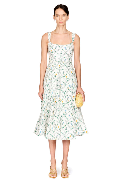 A model stands against a white studio background wearing a sleeveless midi dress with a white base and a green and yellow botanical pattern. She is holding a small yellow clutch and wearing gold sandals.