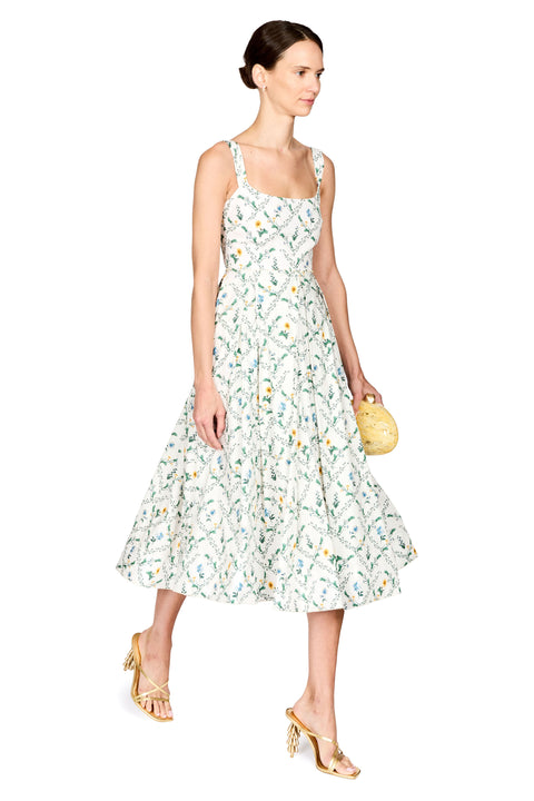 A woman modeling a sleeveless midi dress with a white base and a delicate blue and yellow floral pattern, paired with gold strappy heels and holding a small yellow clutch against a plain white background.