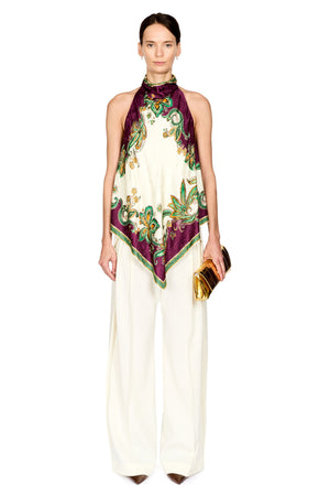 A model wearing a high-neck purple and green ornate patterned silk scarf and white cream pants holding a gold clutch against a plain white background.