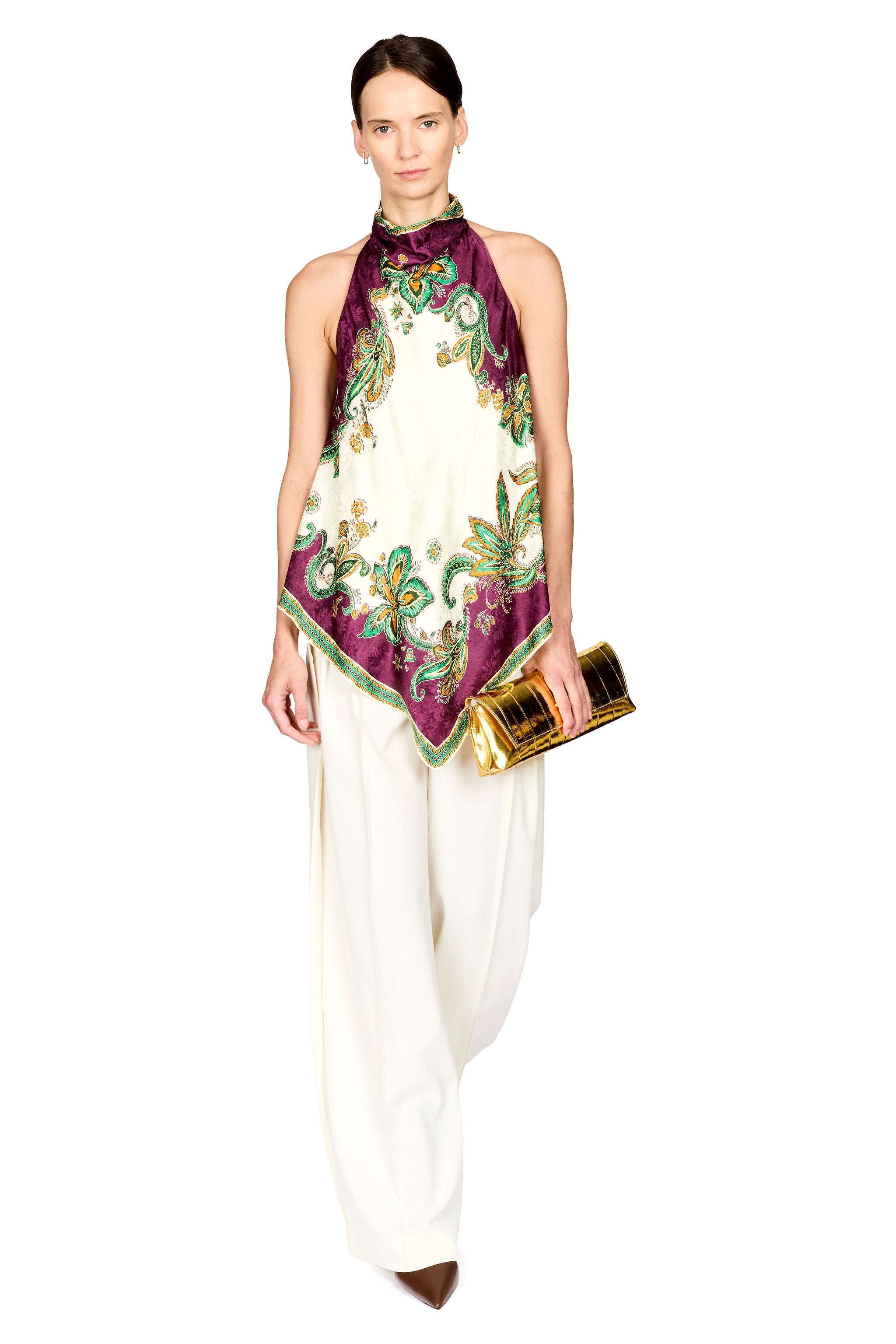 A model wearing a sleeveless cream top with a purple, green, and gold patterned scarf tied around the neck, paired with cream wide-leg pants and holding a gold clutch, set against a plain white background.