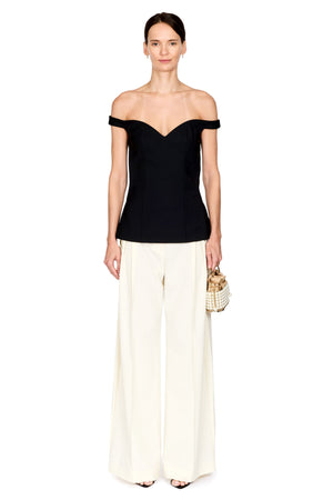 A model stands against a plain white background wearing a black off-the-shoulder top, off-white wide-leg trousers, and holding a small woven handbag.