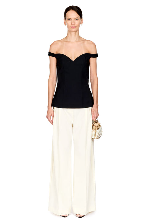 A model stands against a plain white background wearing a black off-the-shoulder top, off-white wide-leg trousers, and holding a small woven handbag.