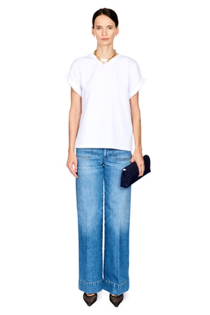 A model stands against a white background wearing a white short-sleeve top, wide-leg blue jeans, black pointed shoes, a gold necklace, and holding a dark blue clutch.