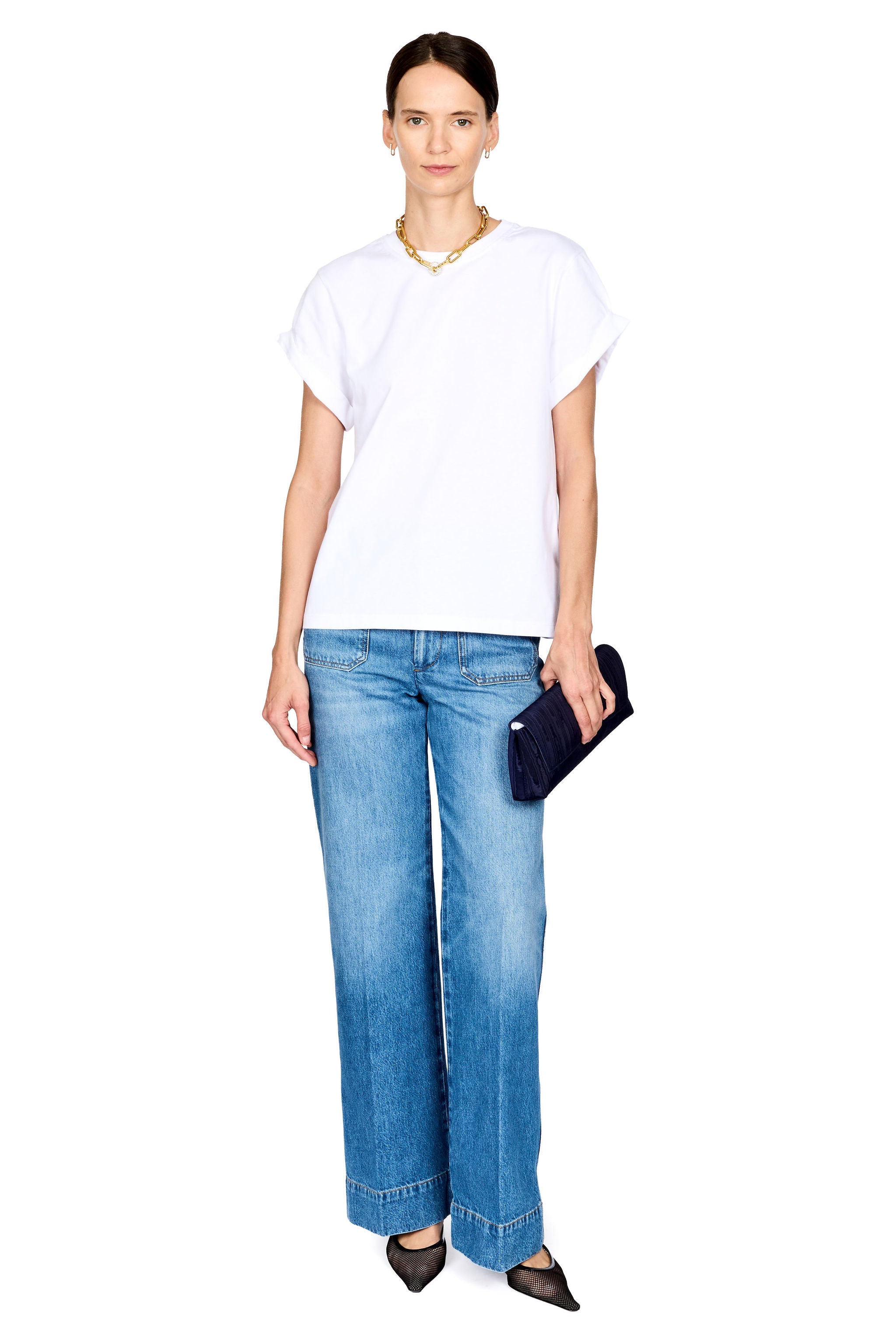 A woman modeling a white short-sleeve t-shirt and wide-leg blue jeans, holding a dark navy clutch purse against a solid white background.