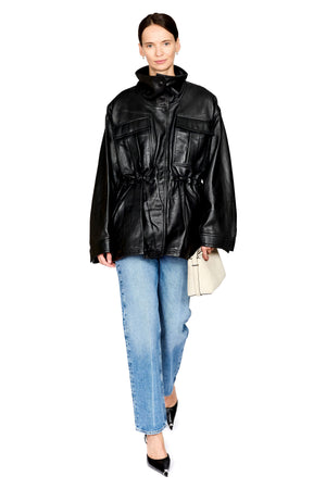 A model wearing an oversized black leather jacket with a high collar and drawstring waist, paired with light blue jeans, black pointed-toe heels, and holding a small cream-colored handbag against a solid white background.