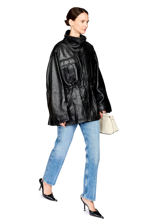 A woman modeling an oversized black leather jacket with a high folded collar, front flap pockets, and a drawstring waist, styled with light blue jeans, black pointed-toe heels, and a small cream handbag against a plain white background.