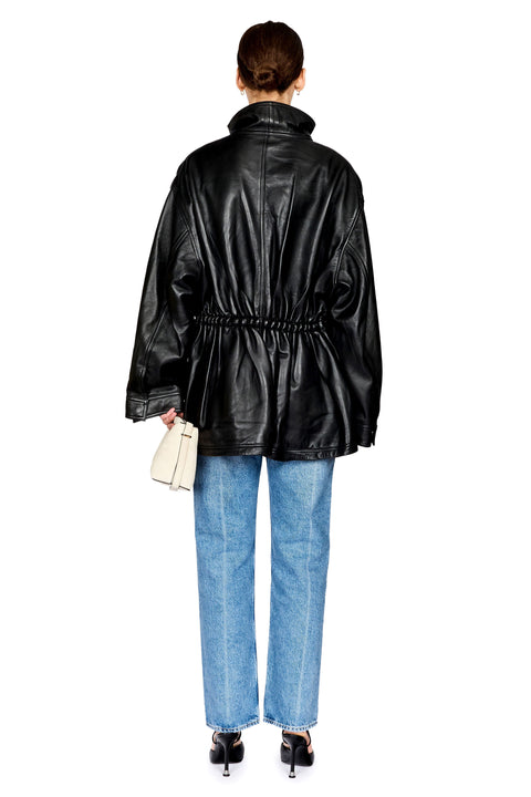 A woman shown from the back wearing an oversized black leather jacket with a drawstring waist, blue jeans, and black heels, holding a small cream clutch against a plain white background.