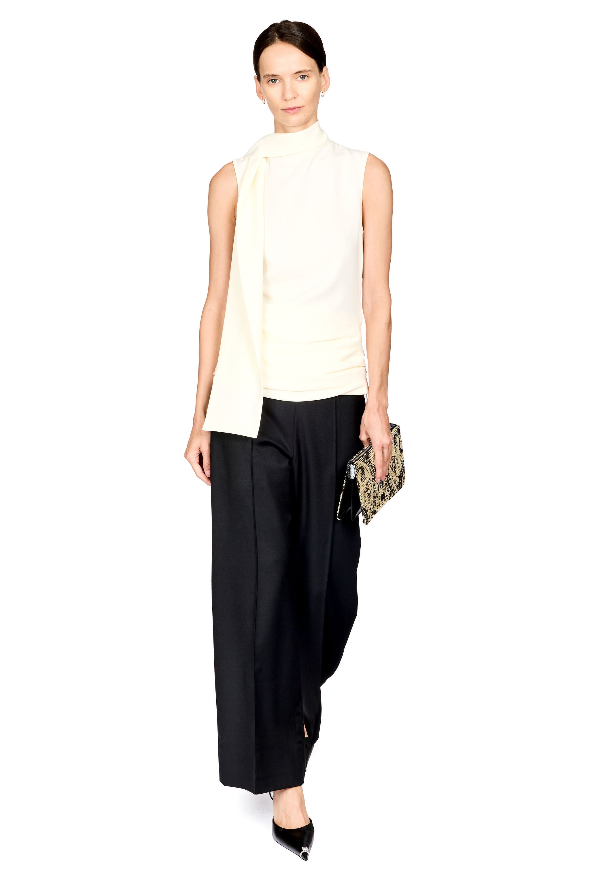 A model wearing a sleeveless cream mock-neck top, black wide-leg draped trousers, and black pointed shoes, holding a small patterned clutch against a plain white background.