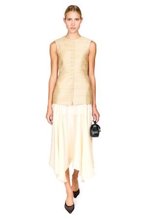 A woman modeling a sleeveless beige textured vest with a button front and a flowing cream high-low skirt, holding a small black handbag against a plain white background.