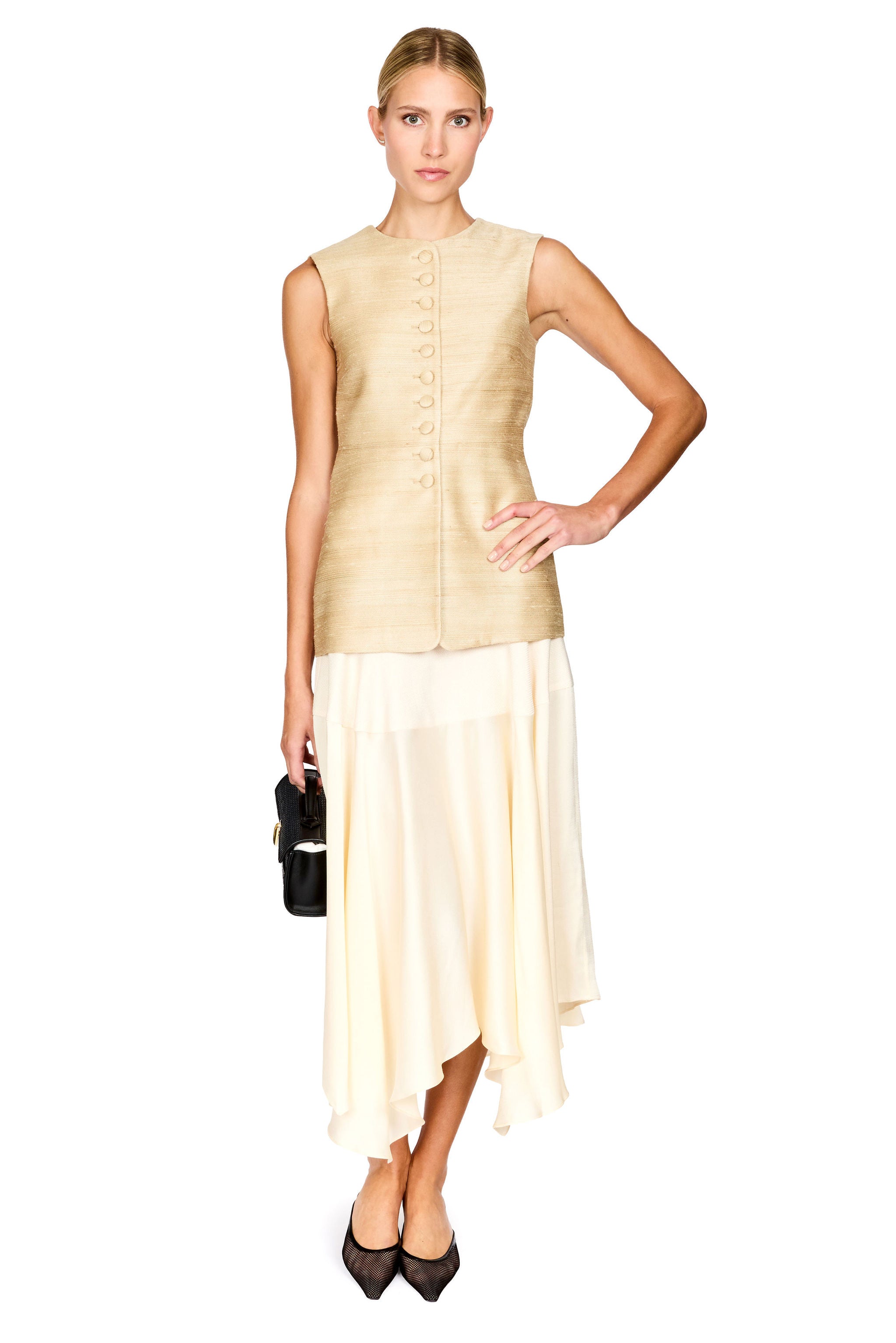 A woman modeling a sleeveless beige textured vest with a button front and a cream asymmetrical midi skirt, holding a small black handbag against a plain white background.
